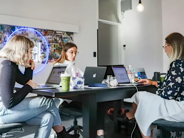 Women working at laptops at a shared desk in a creative office, collaborating on listings for City Directory Base